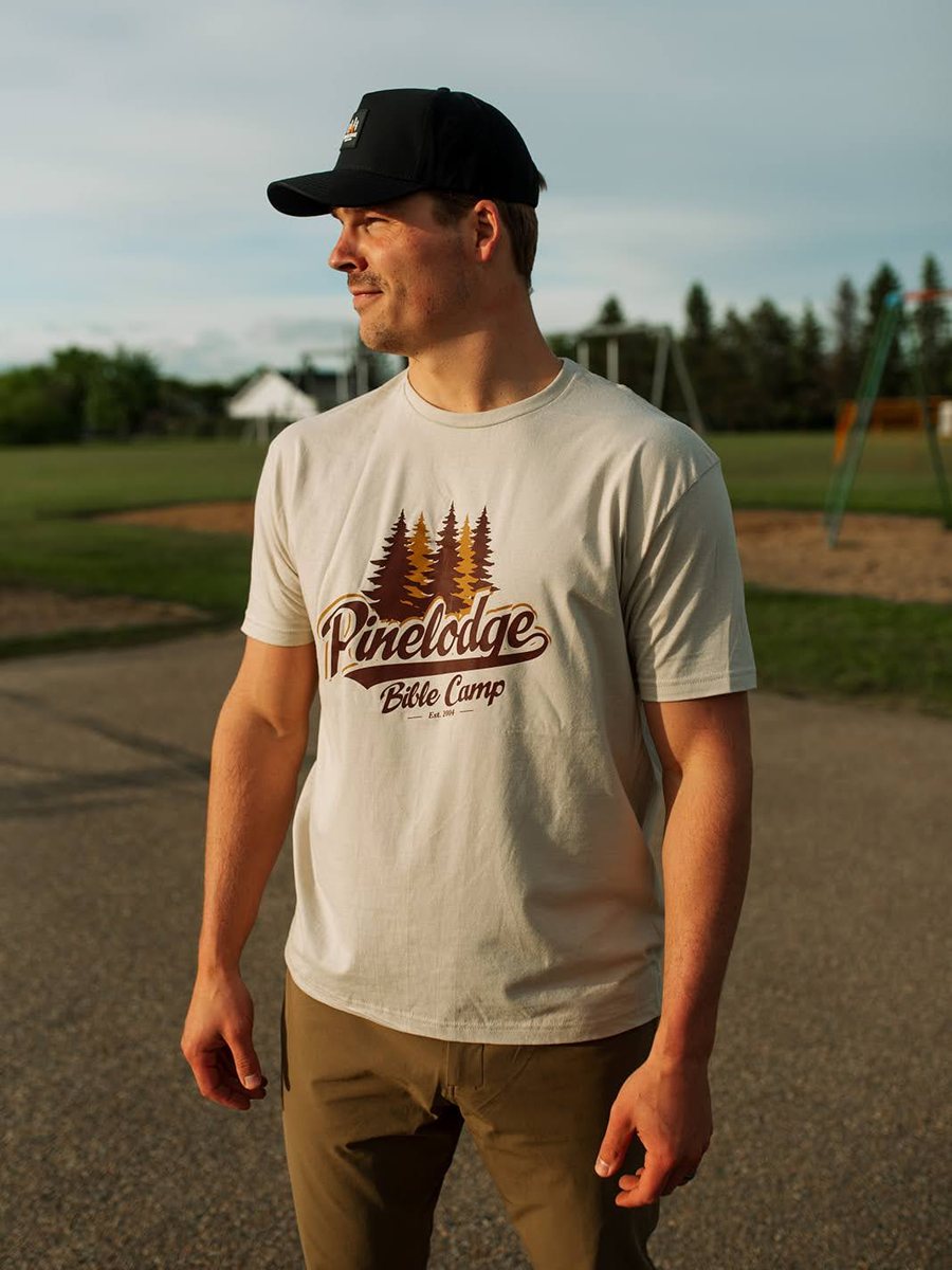 Man in a Pinelodge Bible Camp script t-shirt and black patch hat at a playground in summer evening light